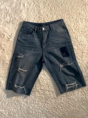 Blå slitna jeansshorts med hål finns också i byxa! - Säljer ett par blå jeansshorts med flera slitningar och hål på framsidan. Shortsen har fem fickor, knappgylf och en relaxed passform som går till knäna. Perfekta för dig som gillar en edgy och avslappnad stil.