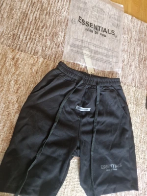 Essentials svart - Snygga svarta shorts från Essentials Fear of God med elastisk midja och långa snören framtill. Shortsen har diskret logga på vänster ben och en liten Essentials-detalj framtill. Perfekta för en avslappnad och trendig stil. Passar även för storlek S