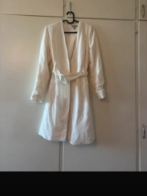 Vit kappa med knytskärp - Stilren vit kappa med knytskärp i midjan och lång ärm. Kappan har en rak passform och öppen front utan synliga knappar. Perfekt för dig som gillar minimalistisk och clean stil.