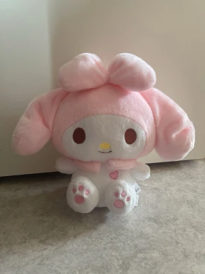 My Melody gosedjur ÄKTA - Supergulligt My Melody gosedjur från Sanrio i mjuk plysch. Den har stora rosa öron, en stor rosett på huvudet och vita tassar med rosa detaljer. Ansiktet är vitt med bruna ögon och gul nos. Perfekt för alla som älskar kawaii och samlar plushies. Den är i nyskick utan defekter med lappen kvar.