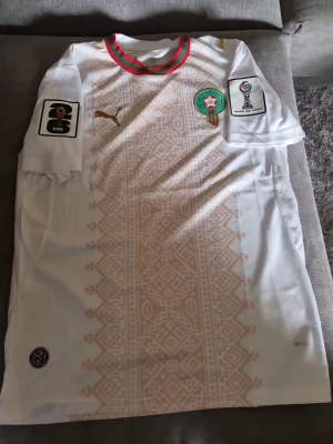 Marocko fotbollströja Puma VM - Snygg vit Marocko fotbollströja från Puma med gulddetaljer och traditionellt mönster framtill. Tröjan har officiella FIFA och VM Qatar 2022-patchar på ärmarna samt Marockos landslagsemblem på bröstet. Röd och grön rand vid kragen. Perfekt för fotbollsfans!