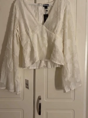 Vit omlottblus från boohoo  - Säljer en vit omlottblus från boohoo med v-ringning och långa, vida ärmar. Blusen har ett diskret blommönster i tyget och lätt transparent material som ger en luftig känsla. Perfekt att styla med jeans eller kjol för en trendig look.