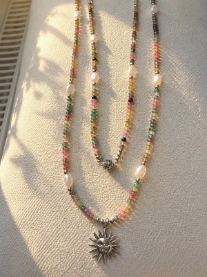 Färgglatt pärlhalsband med solhänge - Unikt halsband med små färgglada glaspärlor och imitationspärlor. Halsbandet har ett detaljerat solformat hänge i rostfritt stål som ger en cool boho-vibe. Perfekt för dig som gillar statement-smycken och vill sticka ut med accessoarer. Magnetlås