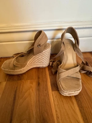 Beiga kilklackade sandaler från Zara - Snygga beiga sandaler från Zara med kilklack och snörning runt ankeln. Skorna har öppen tå, korsade remmar framtill och en flätad sula i espadrillestil. Perfekta för sommaren och ger en trendig look till din outfit. Klackhöjden är 11cm och endast använda en gång! OBS: FRAKTAR INOM 24H!🩷 