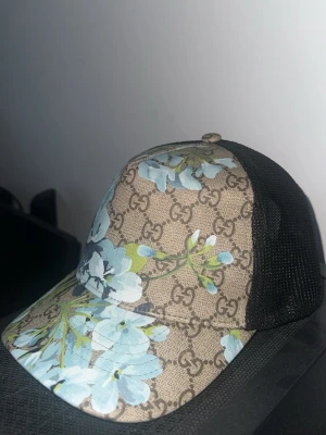 GG  blommig keps  - Säljer en riktigt snygg Gucci-inspirerad truckerkeps med klassiskt monogrammönster och blommigt print i ljusblått. Mesh på sidorna och baktill ger en skön och luftig passform – perfekt till sommaren eller som en stilren detalj i vardagsoutfiten.  ✨ Bra skick ✨ Justerbar passform ✨ Trendig truckermodell ✨ Färger: beige, svart, ljusblå & grön