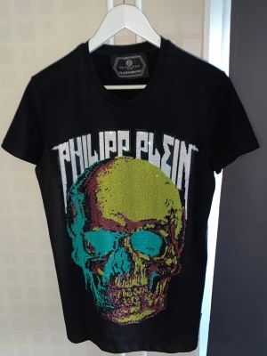 Philipp Plein T-Shirt – Skalle med pärlor – Som ny - Exklusiv Philipp Plein T-shirt med färgglatt skallemotiv och dekorativa pärlor. Storlek S, passar även M. Endast använd en gång och är i mycket fint skick. Originaltaggar finns kvar. En premiummodell som kostade cirka 8 000 kr vid köp år 2021. ✔ Använd endast en gång ✔ Originaltaggar kvar ✔ Pärlor och detaljer intakta ✔ Inga fläckar eller skador ✔ 100 % äkta Perfekt för dig som vill ha en exklusiv designer-T-shirt till en bråkdel av nypriset.