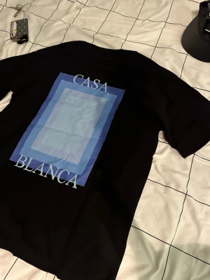 Svart Casa Blanca t-shirt med tryck - Snygg svart t-shirt från Casa Blanca med stort grafiskt tryck i blå toner och texten 'CASA BLANCA' på ryggen samt mindre tryck fram. Klassisk passform och rund hals. Perfekt för dig som gillar streetwear och stilrena detaljer.