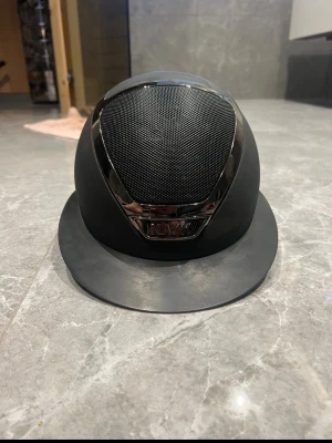 Svart ridhjälm från KASK - Snygg svart ridhjälm från KASK med matt finish och ventilerande mesh framtill. Hjälmen har en elegant design med kromade detaljer och justerbart hakband. Perfekt för dig som vill ha både stil och funktion i stallet. Lite repor, aldrig tappad, storlek 55