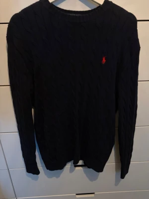 Mörkblå kabelstickad tröja Polo Ralph Lauren - Kabelstickad mörkblå tröja från Polo Ralph Lauren med rund halsringning och den klassiska röda loggan broderad på bröstet. Tröjan har långa ärmar och ribbade muddar. Perfekt för lager-på-lager och snygg till jeans.