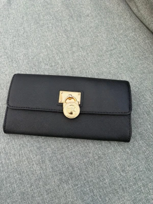 Svart plånbok från Michael Kors - Stilren svart plånbok från Michael Kors i saffianoläder med guldfärgad metallogo och dekorativt lås framtill. Plånboken har flera kortfack, sedelfack och ett myntfack med dragkedja. Perfekt för dig som vill ha både funktion och design.