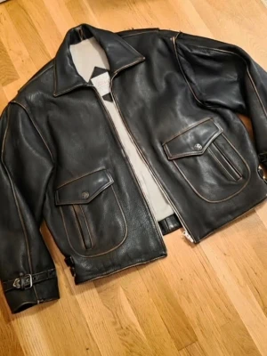 Svart vintage skinnjacka med fickor - Snygg svart skinnjacka med bred krage, två stora fickor framtill med lock och knappar samt justerbara spännen vid ärmsluten. Jackan har en rak passform och dragkedja framtill. Perfekt för dig som gillar en klassisk men cool look.