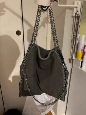 Grå kedjeväska  - Snygg grå handväska med silverfärgade kedjedetaljer och axelremmar. Väskan har en mjuk form och är gjord i läder med dekorativa sömmar längs kanterna. Perfekt för dig som gillar en trendig stil. Använd enstaka gånger, får plats med mycket!💗