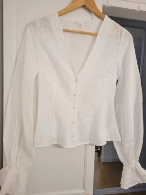 Vit broderad blus från NA-KD - Söt vit blus från NA-KD med broderade blommor, v-ringning och knappar framtill. Blusen har långa puffärmar med resår vid ärmslut och är gjord i ett tunt, luftigt material som känns riktigt somrigt och fräscht.
