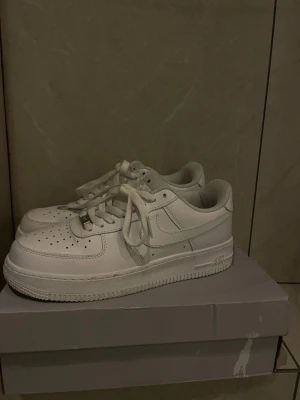 Nike Air Force 1 vita sneakers - Klassiska vita Nike Air Force 1 sneakers i lågt utförande. Skorna har en stilren design med perforerad tå, vita skosnören och den ikoniska swoosh-loggan på sidan. Tillverkade i läder med en rejäl sula för skön komfort.