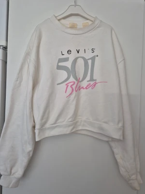 Sweatshirt - Vit sweatshirt från Levi's med stort tryck '501 Blues' i grått och rosa framtill. Tröjan har rund halsringning, ribbade muddar och avslappnad passform. Tillverkad i mjukt bomullsmaterial, perfekt för en chill och stilren look. Finns fläckar.