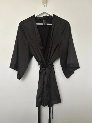 Svart Kimono - H&M - Stilren svart morgonrock i kimonomodell från H&M. Tillverkad i ett glansigt, mjukt material med knytskärp i midjan och vida trekvartsärmar. Använd och i fint skick. Storlek S. 
