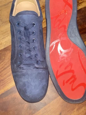 Mörkblå sneakers från Christian Louboutin - Snygga mörkblå sneakers i mocka från Christian Louboutin med klassisk röd sula och diskret logga undertill. Skorna har rund tå, matchande blå skosnören och metallöljetter. Perfekta för dig som vill ha en exklusiv och stilren look.