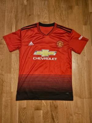 Manchester United matchtröja Adidas - Röd Manchester United matchtröja från Adidas med svarta detaljer och klubbmärke på bröstet. Tröjan har korta ärmar, V-ringning och sponsorlogga från Chevrolet på framsidan. Tillverkad i lätt och ventilerande material, perfekt för fotbollsträning.