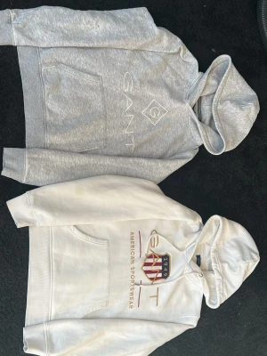 Två GANT hoodies med tryck - Båda passar 164 Säljer två snygga hoodies från GANT i storlek 158/164. En är ljusgrå med vitt GANT-tryck och en är vit med GANT-logga och American Sportswear-tryck i rött och guld. Båda har huva och magficka, perfekta för en avslappnad stil.
