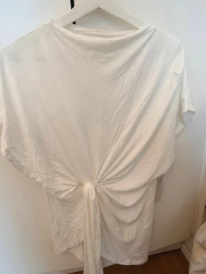 Vit topp med knyt i midjan - Säljer en vit topp med snygg knytning i midjan som ger en cool draperad effekt. Toppen har en lös passform och är tillverkad i mjukt bomullsmaterial. Perfekt för dig som gillar enkla men ändå unika detaljer.