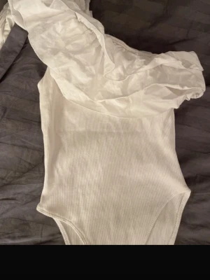 Zara volang body - Säljer en snygg vit body med ribbad struktur och off-shoulder design. Bodyn har en volangdetalj över axeln. Perfekt för sommaren. Storlek S men passar även xs. 
