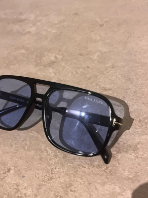 Svarta Tom Ford solglasögon - Snygga svarta solglasögon från Tom Ford med fyrkantig form och blåtonade glas. Bågarna är i blankt svart material med en cool detalj i metall vid gångjärnen. Perfekta för att lyfta din stil med en exklusiv touch.