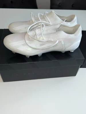 Vita Adidas fotbollsskor - Säljer ett par stilrena vita fotbollsskor från Adidas med låg vikt och modern design. Skorna har snörning, mönstrad ovandel i syntetmaterial och fasta dobbar för bra grepp på planen. Perfekta för dig som vill ha både stil och funktion på fotbollsplanen. Årsmodellen är 2023 