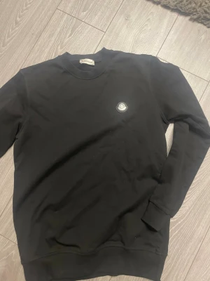 Svart sweatshirt från Moncler - Stilren svart sweatshirt från Moncler med rund halsringning och diskret Moncler-logga på bröstet. Tröjan har långa ärmar och ribbade muddar vid ärmslut och nederkant. Perfekt för en clean och avslappnad look.