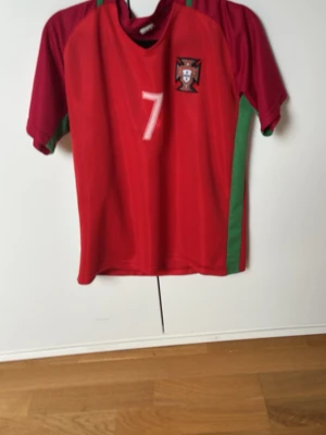 Portugal Ronaldo matchtröja röd - Säljer en röd Portugal fotbollströja med gröna detaljer och nummer 7 på bröstet och ryggen. På ryggen står det 'Ronaldo'. Tröjan har korta ärmar och Portugal-emblemet på framsidan. Perfekt för dig som älskar fotboll och Cristiano Ronaldo.
