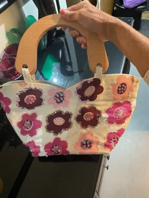 Blommig handväska med trähandtag - Unik handväska i tyg med stora trähandtag och färgglada blommor i rosa, lila och vinrött på framsidan. Väskan har en vit bas och dekorativa paljetter på blommorna för extra bling. Perfekt accessoar för dig som gillar retro och statement-väskor.har en inne ficka med dragkedja och man stänger väska med dragkedja. Det finns missfärgning på kanten av väskan ,som man ser på bilden 5och sexan .men det syns inte