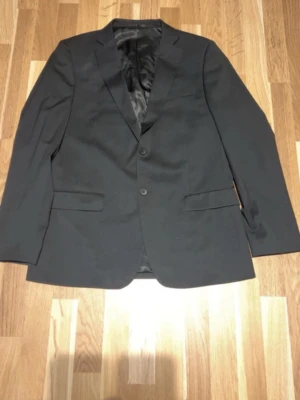 Svart klassisk kavaj med två knappar - Stilren svart kavaj med två knappar framtill och klassiska fickor. Kavajen har smal passform, helfodrad insida och fyra knappar vid ärmsluten. Perfekt till skjorta och byxor för en dressad look.
