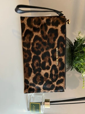 Leopardmönstrad kuvertväska Michael Kors - Snygg kuvertväska från Michael Kors i leopardmönstrad mockaimitation. Väskan har guldfärgade detaljer, dragkedja upptill och en avtagbar handledsrem. Perfekt för dig som vill sticka ut med ett djurmönster och gillar trendiga accessoarer. Aldrig använt, lappen kvar.