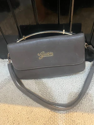 Grå axelväska från Guess - Snygg grå axelväska från Guess med guldfärgad logga på framsidan. Väskan har ett stilrent och klassiskt rektangulärt format, justerbar axelrem och ett metallhandtag upptill. Materialet är slätt konstläder som ger en elegant look.