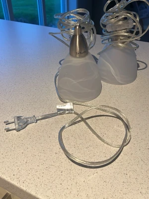 Två vita taklampor i glas - Säljer två stilrena taklampor i vitt glas med mjukt mönster och transparent sladd. Lamporna har konformad skärm och detaljer i metall upptill. Perfekta för att skapa mysig belysning i hemmet.