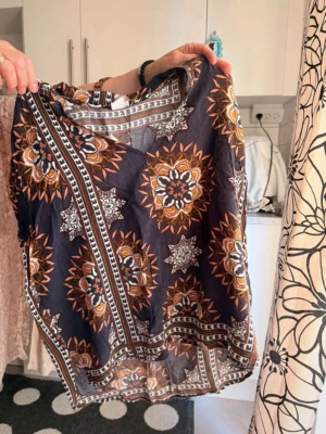 Mönstrad blus i boho-stil - Säljer en luftig blus med mörkblå bas och stora, bohemiska mönster i orange, vitt och brunt. Blusen har v-ringning och korta ärmar, perfekt för dig som gillar färgstarka och unika plagg. Materialet känns lätt och skönt mot huden.
