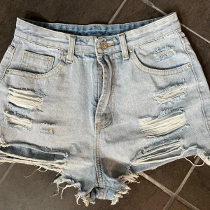 Ljusa denimshorts med slitningar - Säljer ett par ljusblå denimshorts med råa kanter och flera slitningar både fram och bak. Klassisk femficksmodell med bälteshällor och knappgylf. Perfekta för en avslappnad och trendig sommarstil.