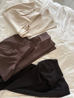 Tre par kostymbyxor i olika färger - Säljer tre par stilrena kostymbyxor i beige, brun och svart. Byxorna har raka ben, pressveck och midjehällor för bälte. Perfekta för en clean och modern look. Materialet känns mjukt och har ett snyggt fall. Köpte från NAKD , aldrig använt, storlek 36, men passar 34 också 