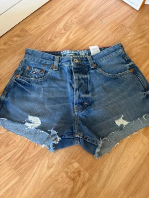 Blå jeansshorts från Hilfiger Denim - Snygga blå jeansshorts från Hilfiger Denim med råa kanter och slitna detaljer. Klassisk femficksmodell med hög midja och avslappnad passform. Perfekta för en chill sommarstil.