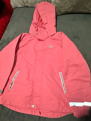 Rosa vindjacka med huva - Snygg rosa vindjacka från Didriksons med huva och dragkedjor på fickorna. Jackan har reflexdetaljer vid ärmslut och en diskret logga på bröstet. Perfekt för blåsiga dagar och enkel att matcha med olika outfits.