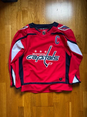 Washington Capitals Ovechkin matchtröja - Säljer en röd Washington Capitals hockeytröja från Adidas med Ovechkin och nummer 8 på ryggen. Tröjan har vita och marinblå detaljer, NHL-märke vid halsen och ett stort Capitals-tryck på bröstet. Långärmad och tillverkad i funktionsmaterial.
