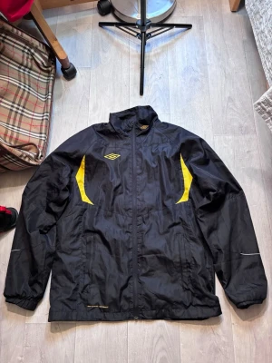 Svart vindjacka från Umbro - Snygg svart vindjacka från Umbro med gula detaljer på bröstet och ryggen. Jackan har hög krage, dragkedja framtill och två sidofickor. Tillverkad i lätt och tåligt syntetmaterial, perfekt för blåsiga dagar. Umbro-logga på bröstet och ryggen.