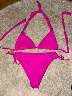 Rosa trekantsbikini med knyt - Snygg rosa trekantsbikini med knyt i sidorna på underdelen och i nacken samt ryggen på överdelen. Klassisk modell som ger en fräsch och ungdomlig look, perfekt för stranden eller poolen. Materialet känns mjukt och följsamt. Aldrig använt!!!