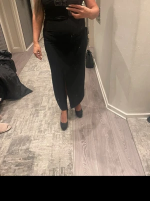 Svart långklänning med slits - Elegant svart långklänning med smala axelband och slits framtill. Klänningen har en snygg knyt-detalj i ryggen och figurnära passform. Perfekt för dig som vill ha en stilren och klassisk look.