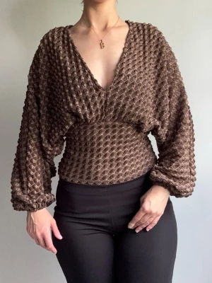 Brun strukturerad blus med v-ringning - Säljer en brun blus med unik struktur och djup v-ringning. Blusen har långärmade, puffiga ärmar och smockad midja som ger en snygg siluett. Materialet är stretchigt och har ett 3D-mönster som gör den extra speciell. Perfekt för dig som gillar trendiga detaljer.