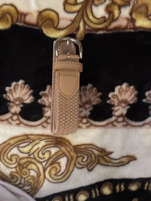 Beige flätat bälte med silverdetalj från Zara - Snyggt beige flätat bälte med silverfärgat spänne och detaljer i skinnimitation. Perfekt för att lyfta vilken outfit som helst och ger en trendig touch till jeans eller byxor.