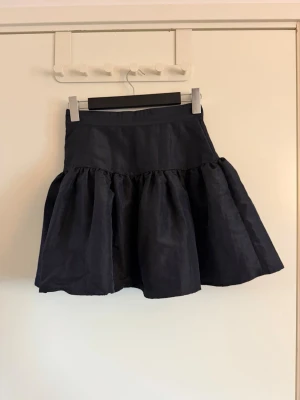 Svart volangkjol med hög midja - Snygg svart kortkjol med volang och hög midja. Kjolen har en fluffig och utställd form som ger en cool siluett. Perfekt att styla med en croppad topp eller stickad tröja. Materialet känns lätt och har en diskret glans.
