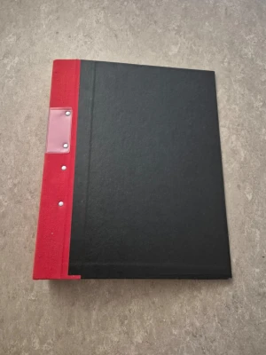 Pärm - Säljer en klassisk pärm från Jopa med svart omslag och röd rygg. Pärmen har metallmekanism för papper och plastficka på ryggen för etikett. Perfekt för att organisera dokument, anteckningar eller samlingar. Tillverkad i hård kartong och tygklädd rygg.