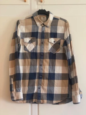 Rutig skjorta från vans - Snygg rutig skjorta från vans i beige, blått och vitt. Skjortan har två bröstfickor med knappar, klassisk krage och långa ärmar. Tillverkad i mjuk bomull och perfekt för en avslappnad stil.