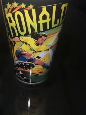 Ronaldinho plastmugg McDonald's FIFA - Cool plastmugg med färgglad tecknad bild av Ronaldinho i brasiliansk landslagströja. Muggen har detaljer i gult, grönt och blått samt texten 'RONALDINHO' och FIFA-logga. Perfekt för fotbollsfans och samlare.
