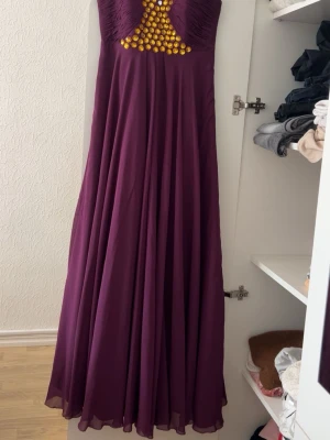Lång lila klänning med gulddetaljer - Elegant långklänning i lila med guldfärgade pärlor framtill vid bysten. Klänningen har en flowy kjol och är ärmlös med rynkade detaljer upptill. Perfekt för speciella tillfällen där du vill sticka ut. Dock så är få paljetter på klänningen av. Klänningen är från Istanbul i Turkiet.